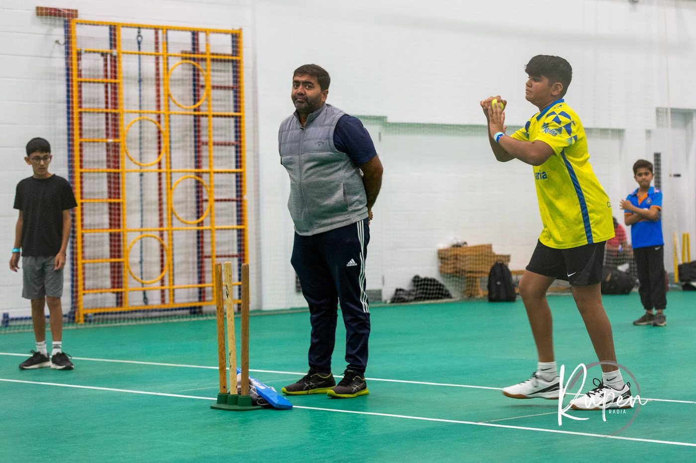 A young player bowling in the indoor cricket hall, stumps set up behind the batter